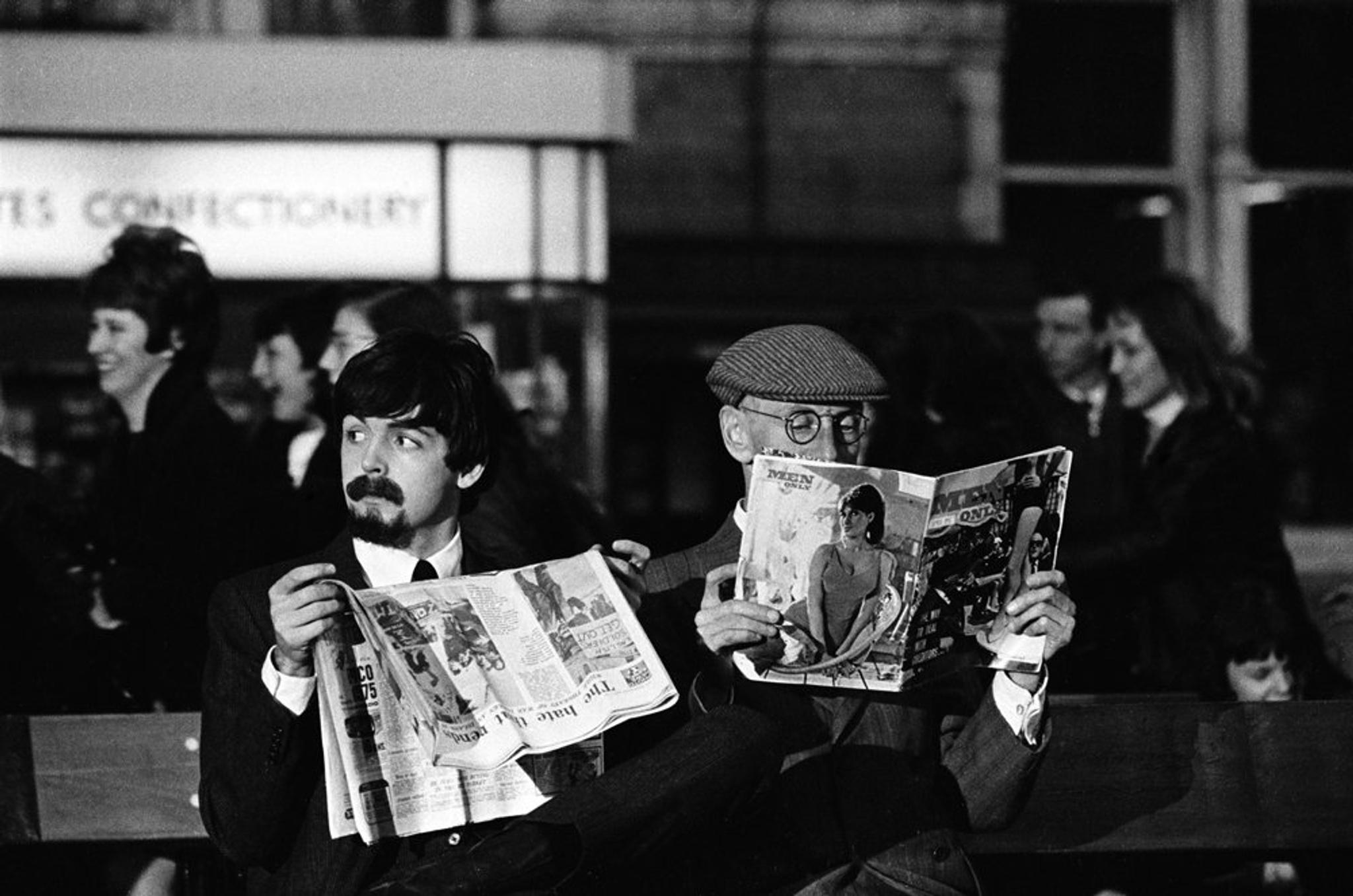 Paul hiding his face behind a newspaper sitting next to Wilfrid Brambell at the train station