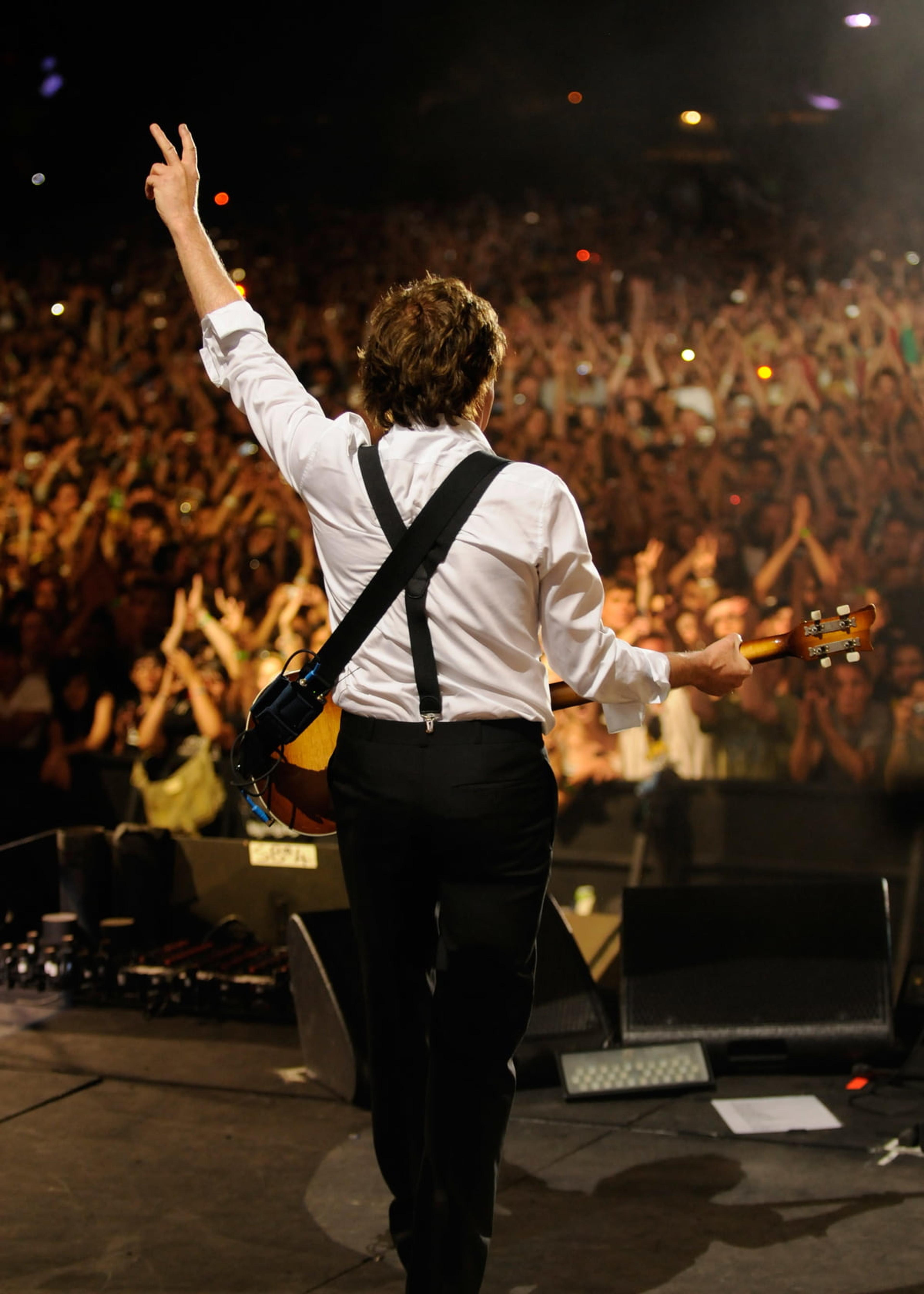 Paul stands facing a crowd and making a peace sign with his left hand
