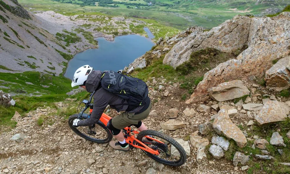 Mountain biker with Evoc backpack riding a rocky descent high in the mountains with a lake in the background