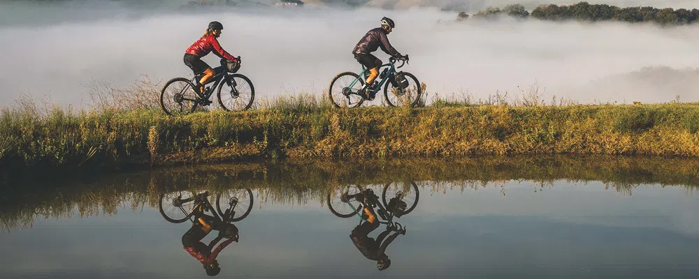 two cyclists on electric gravel bikes pass a mountain lake with cloud behind