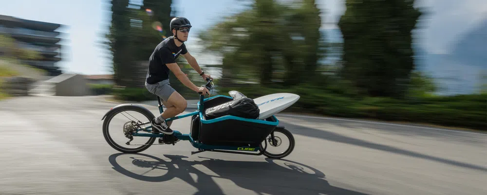 A man rides past on an electric cargo bike holding a surfboard