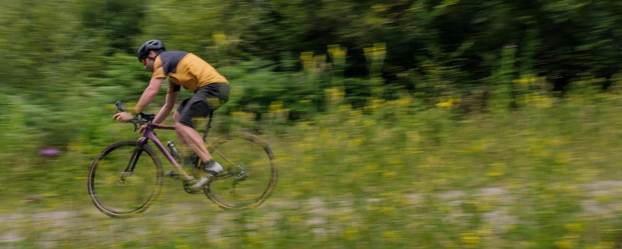 Gravel cyclist riding fast downhill in open woodland past yellow flower undergrowth