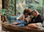 Two female friends curled on a sofa together looking at a phone and smiling. They're in front of a window with lots of trees outside