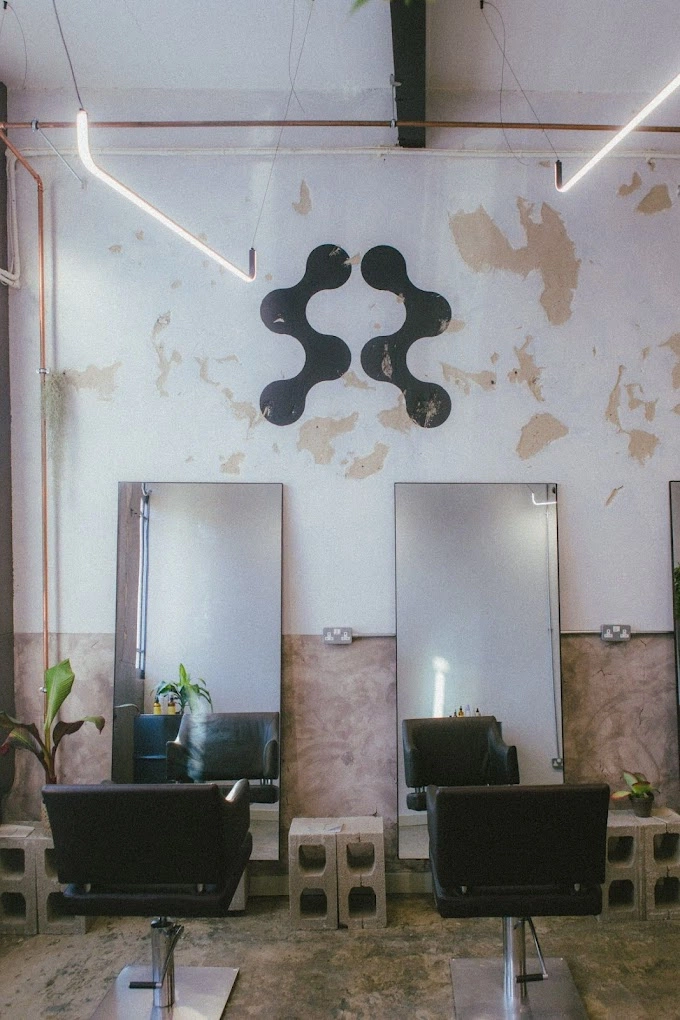 This image shows the APRE logo proudly positioned on the salon wall. Minimalist interior design, concrete floors, distressed walls, two black salon chairs in front of large mirrors, exposed copper pipes and industrial lighting fixtures.