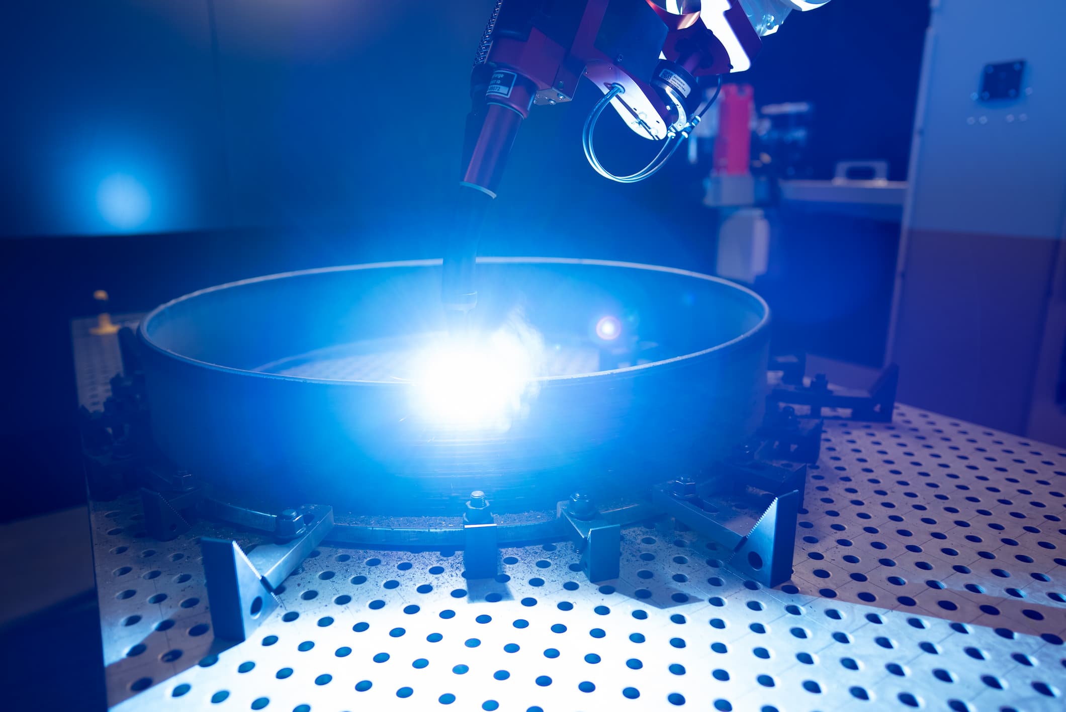 WAAM in print with blue sparks coming from a robot arm as it forms a metal cylinder. 