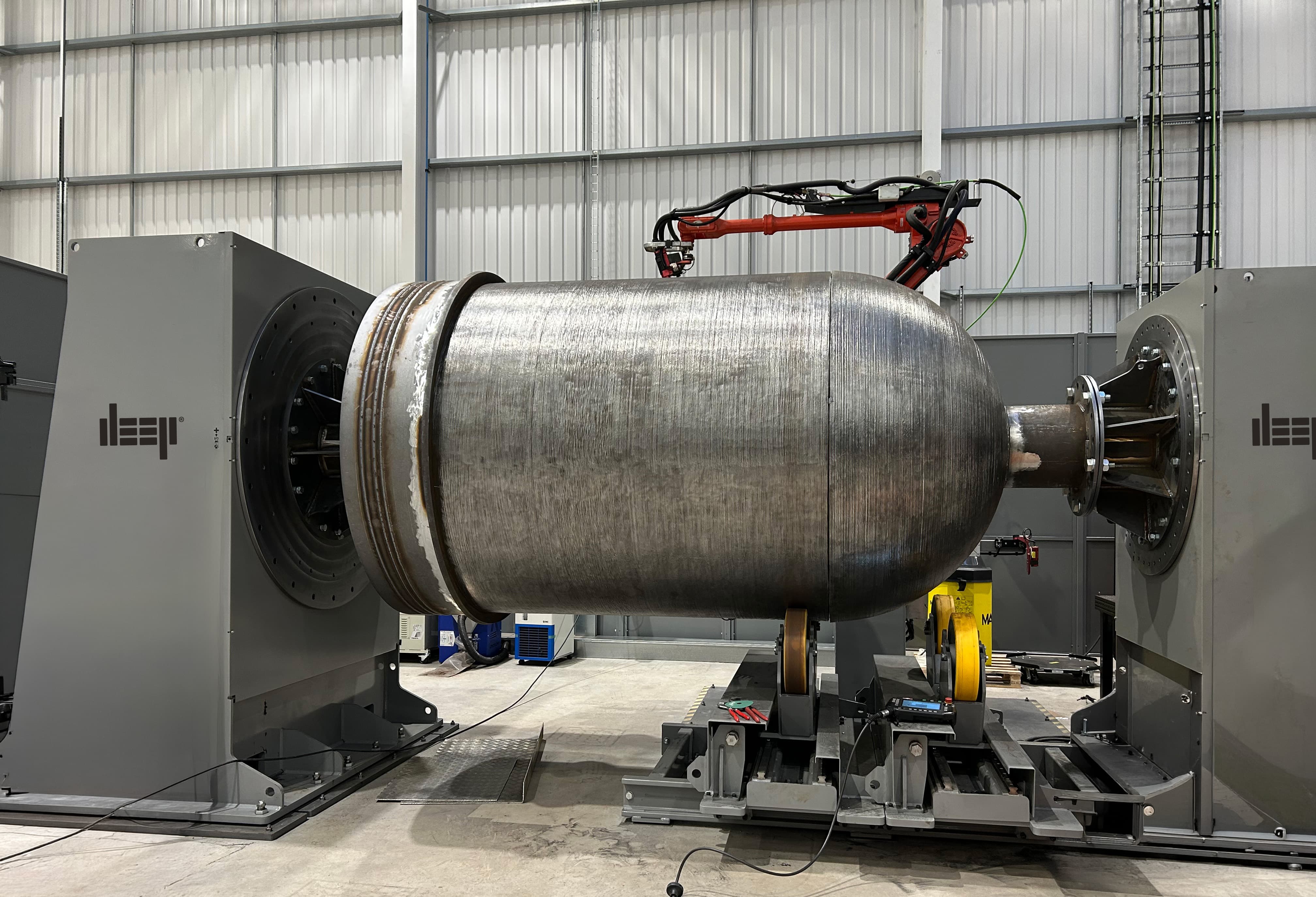 A two axis manipulator WAAM robot inside a large warehouse with a printed hemisphere to the left of the image. 