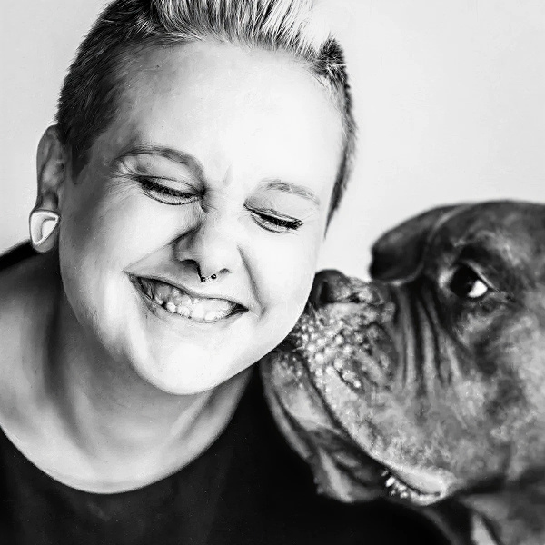 Black-and-white photo of Carolin Jütz. A Boxer-Mix dog kissing her face while she laughs.