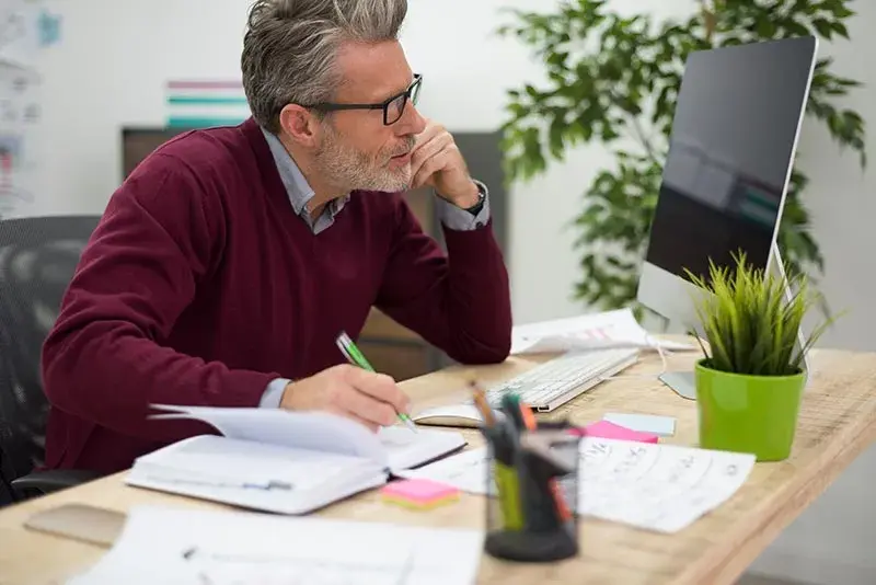 Manager con aria pensierosa davanti al computer e all'agenda, immagine che rappresenta la riflessione e la pianificazione come competenze soft fondamentali per un imprenditore.